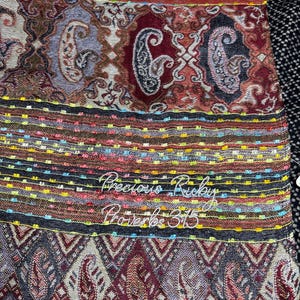 May include: A woven textile featuring a multi-colored paisley and geometric pattern in shades of red, brown, and gold. The fabric includes horizontal bands with embroidered text that reads Precious Ruby Proverbs 3:15 in white cursive script.