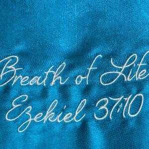 Può includere: Primo piano di un tessuto turchese con testo ricamato bianco. Il testo recita "Breath of Life" e "Ezekiel 37:10" in un carattere corsivo. Il tessuto ha un aspetto strutturato.