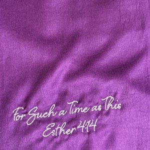 May include: A deep purple fabric with the embroidered text "For Such a Time as This Esther 4:14" in white script. The fabric has a subtle sheen and visible texture, with soft folds and shadows.