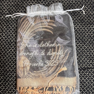 May include: A black and gold scarf with a feather design, packaged in a sheer white drawstring bag. The scarf has a gold fringe and the text "She is clothed in strength & dignity Proverbs 31:25" in white script.