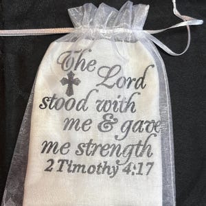 Op de afbeelding: Een witte stoffen tas met de tekst "The Lord stood with me & gave me strength 2 Timothy 4:17" in grijs schrift. Een grijs kruis staat boven de tekst. De tas zit in een doorzichtige witte trekkoordzak.