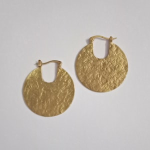 May include: A pair of gold-colored hoop earrings. Each earring has a textured, hammered surface and a circular design with a curved opening. The earrings are displayed on a white background.