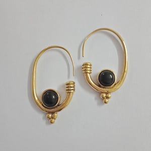 Può includere: Un paio di orecchini a cerchio color oro con una pietra nera al centro di ciascuno. Gli orecchini hanno un design unico con un piccolo gruppo di tre perline in basso.