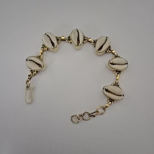 Może przedstawiać: Złota bransoletka z łańcuszkiem i białymi muszelkami kauri. Bransoletka ma zapięcie na karabińczyk.