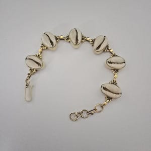 May include: A gold-toned metal chain bracelet featuring five off-white cowrie shells spaced evenly along the links. The bracelet includes a small lobster claw clasp and an adjustable extender chain with circular links at one end for a secure fit.