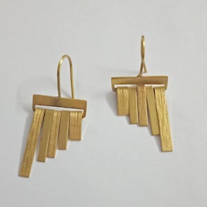 May include: A pair of gold-coloured earrings. Each earring has a rectangular top with a curved hook and several vertical, textured rectangular bars hanging below. The earrings are shown against a white background.