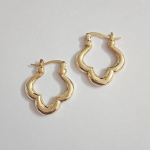 May include: A pair of gold-colored hoop earrings. Each earring features a quatrefoil design, with a hinged closure. The earrings are displayed on a white background, showcasing their polished, reflective surface.