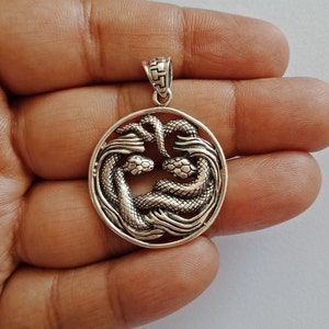 以下が含まれることがあります： 2匹の蛇が絡み合い、詳細な鱗で円形のデザインを形成したシルバーペンダント。ペンダントの上部にチェーンに取り付けるためのループがあります。