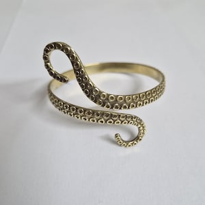 Op de afbeelding: Een messingkleurige armmanchet met een ontwerp van een octopus-tentakel. De manchet wikkelt zich om de arm met een band en een gebogen tentakel. De tentakel heeft gedetailleerde zuignappen. De sieraden liggen op een witte ondergrond.