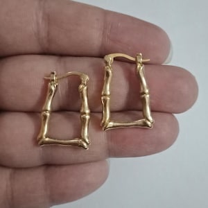 Może przedstawiać: Para złotych, prostokątnych kolczyków koła. Kolczyki mają bambusowy wzór, z teksturowaną powierzchnią i zapięciem na zawias. Kolczyki są prezentowane na neutralnym tle.