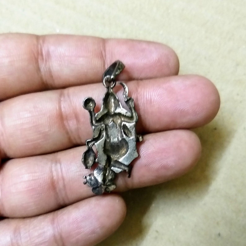 May include: A silver pendant depicting a detailed figure with multiple arms, holding various objects. The pendant has a loop at the top for a chain, possibly a religious or symbolic item. The design is intricate and shows signs of wear.