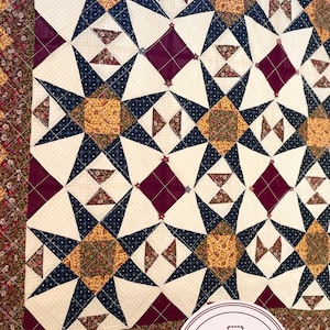 Könnte beinhalten: Eine Steppdecke mit einem sich wiederholenden Muster aus dunkelblauen, burgunderroten und goldenen Sternen auf weißem Hintergrund. Die Steppdecke ist mit einem burgunderroten, goldenen und floral gemusterten Stoff eingefasst. Die Steppdecke ist mit "Patchworkangel(A) Alles aus Textil" beschriftet.