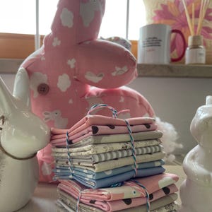 Könnte beinhalten: Ein Stapel Stoffquadrate in Rosa, Blau, Weiß und Braun mit weißen Punkten, Streifen und Blumenmustern. Der Stoff ist mit Schnur zusammengebunden. Der Stoff liegt auf einem Tisch neben einem rosa-weißen Stoffhasen.