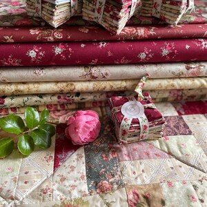Könnte beinhalten: Ein Stapel Stoffbündel in verschiedenen Rot-, Creme- und Grüntönen mit floralen Mustern. Die Bündel sind mit weißem Band gebunden und haben ein weißes Etikett mit einem floralen Design. Der Stoff ist auf einem Patchwork-Steppbett in Creme-, Rot- und Grüntönen angeordnet.