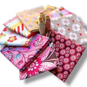 Könnte beinhalten: Eine Sammlung bunter Stoffquadrate mit Blumen- und Mustermotiven, darunter Rosa, Gelb und Blau. Drei Garnrollen in neutralen Farbtönen sind ebenfalls zu sehen, was auf ein Näh- oder Bastelthema hindeutet.