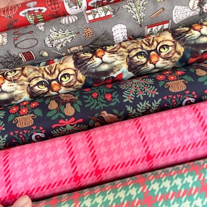 Könnte beinhalten: Ein Stapel aus fünf Stoffballen mit verschiedenen Mustern, darunter Rot mit Feiertagsmotiven, Grau mit Wintersymbolen, ein Druck von Katzen mit Brillen, ein dunkelblaues Blumenmuster sowie zwei pinke und grüne Hahnentritt-Karomuster.