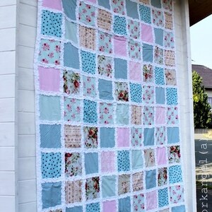Könnte beinhalten: Eine Patchwork-Decke mit verschiedenen Blumenmustern in Rosa-, Blau- und Weißtönen. Die Decke besteht aus Quadraten und ist mit weißer Spitze eingefasst.