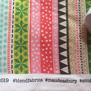 Könnte beinhalten: Nahaufnahme eines weißen Stoffes mit einem bunten Streifenmuster. Der Stoff zeigt geometrische Formen, darunter Dreiecke, Sterne und Punkte. Der Stoff ist mit dem Text "#blendfabrics #maudeasbury #snuglysweater 4. © 2019" beschriftet.