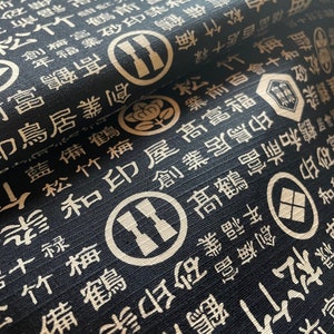 Könnte beinhalten: Schwarzer Stoff mit einem sich wiederholenden Muster aus weißen japanischen Schriftzeichen und Symbolen. Der Stoff hat eine strukturierte Oberfläche.