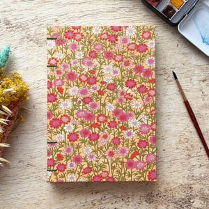 Könnte beinhalten: Ein rechteckiges Notizbuch mit gelbem Einband, das mit einem dichten Muster aus rosa, roten und weißen Blumen auf grünen Stielen bedruckt ist. Das Notizbuch ist am linken Rand mit grünem Faden gebunden und liegt neben einem Pinsel und Trockenblumen.