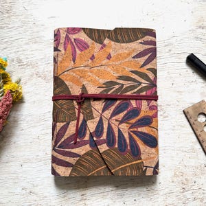 May include: A cork journal featuring a botanical leaf design in orange, purple, and green hues. It's secured with a burgundy elastic band. A black pen and a wooden ruler are also present.
