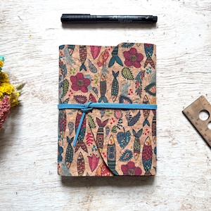 May include: A cork journal with a colorful fish pattern, secured with a blue elastic band. A black pen and a wooden ruler are also visible. The journal's design includes flowers and paisley shapes.