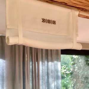 May include: White roman blind with a simple embroidered design. The blind is rolled up and hanging from a wooden beam. The blind is in front of a window with grey curtains and a view of green trees.