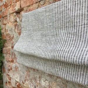 May include: A white linen fabric with thin, dark grey stripes. The fabric is folded in half and draped over a brick wall.