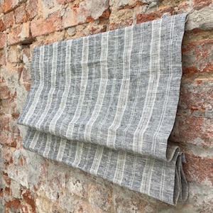 May include: A grey and white striped linen roman blind hanging on a brick wall. The blind is made from a natural fabric with a subtle texture.