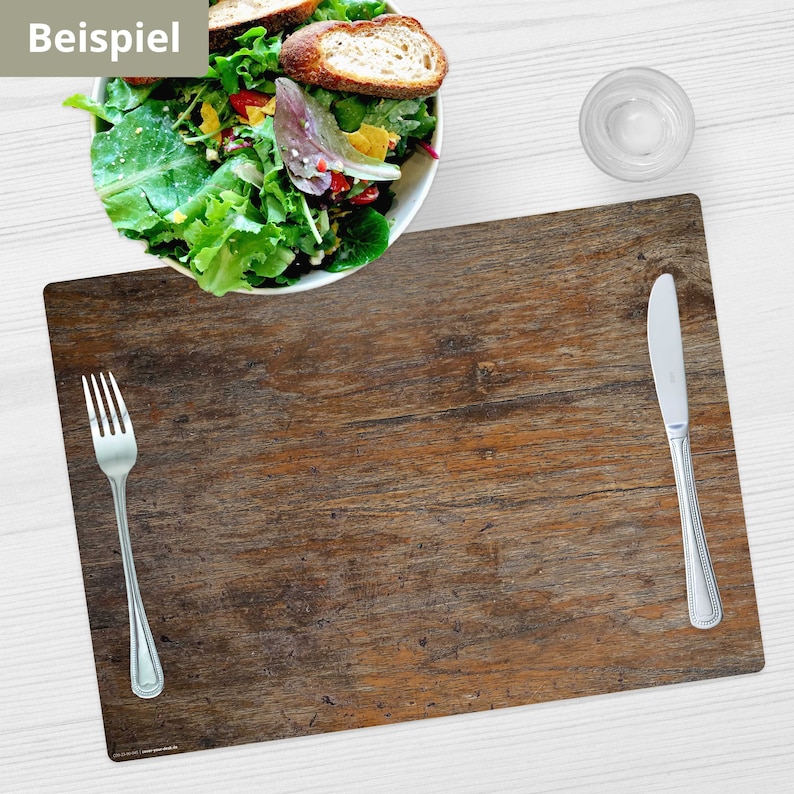 May include: A brown wooden placemat with a rustic wood grain pattern. A silver fork and knife are placed on the placemat. The placemat is on a white wooden table.
