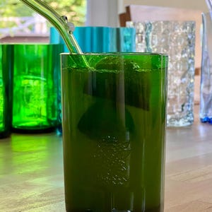 May include: A green glass tumbler with a clear glass straw, filled with a green liquid and ice cubes. Other green and blue glass tumblers are in the background. The glass has a textured surface.
