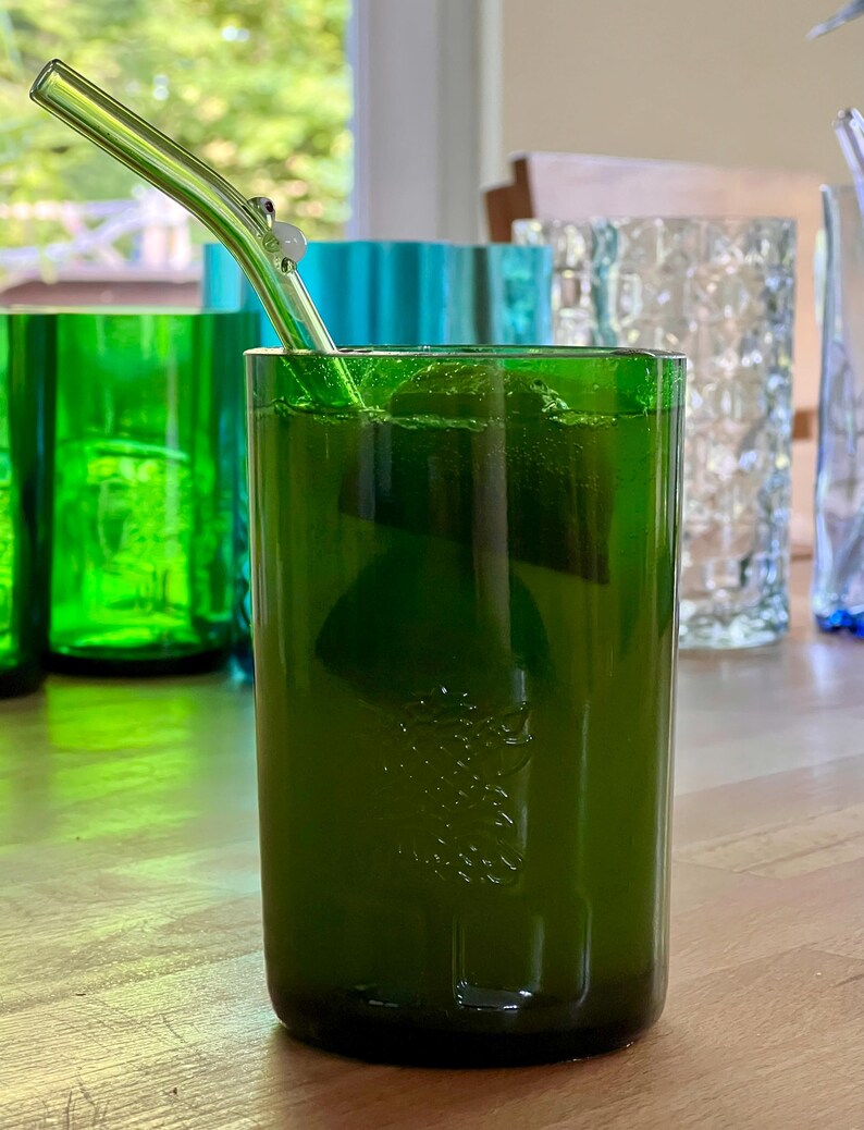 May include: A green glass tumbler with a clear glass straw, filled with a green liquid and ice cubes. Other green and blue glass tumblers are in the background. The glass has a textured surface.
