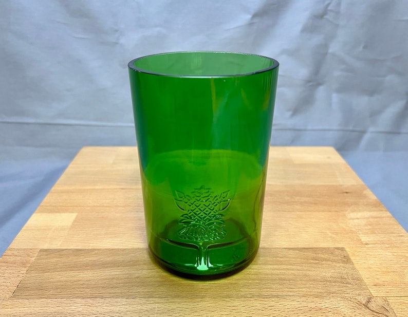 May include: A green glass tumbler with a decorative tree design etched near the base. The glass is cylindrical with a slightly rounded rim. The glass is made of a transparent green material.