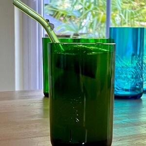 May include: A green glass with a metal straw, filled with a green liquid, sits on a wooden table. The glass is cylindrical and has a dark green hue. Other green and blue glasses are visible in the background, near a window.