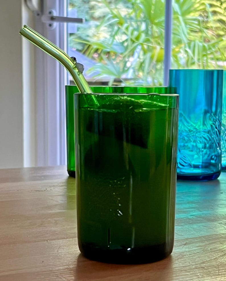 May include: A green glass with a metal straw, filled with a green liquid, sits on a wooden table. The glass is cylindrical and has a dark green hue. Other green and blue glasses are visible in the background, near a window.