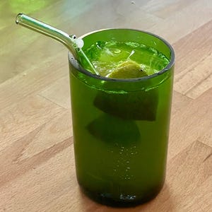 May include: A green glass with a drink inside, featuring a clear glass straw. The drink contains ice and a lime wedge. The glass is sitting on a wooden surface.