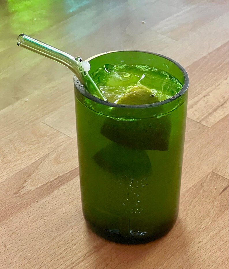 May include: A green glass with a drink inside, featuring a clear glass straw. The drink contains ice and a lime wedge. The glass is sitting on a wooden surface.