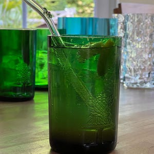 May include: A green glass with a drink and a clear glass straw. The glass has a tree design etched into the side. Other green and blue glasses are in the background. The drink has a lime wedge and ice.