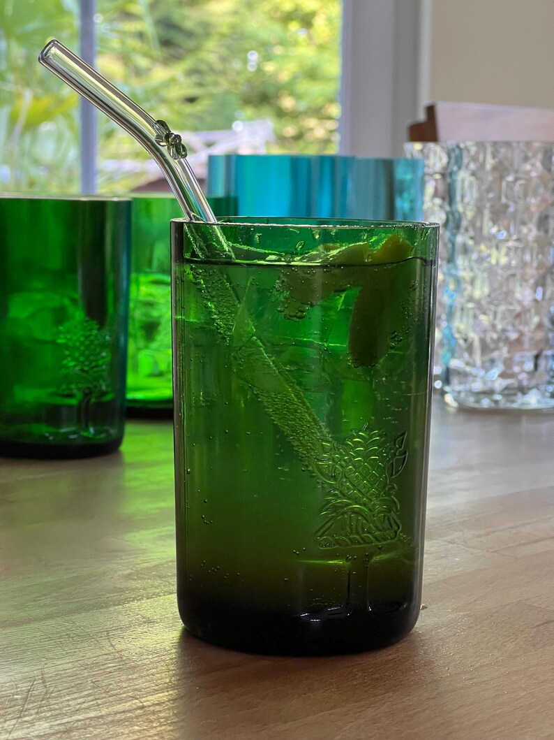 May include: A green glass with a drink and a clear glass straw. The glass has a tree design etched into the side. Other green and blue glasses are in the background. The drink has a lime wedge and ice.