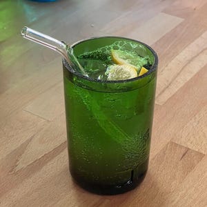May include: A green glass tumbler filled with a green beverage, ice, and lemon slices. A clear glass straw extends from the drink. The glass has a textured surface and is sitting on a wooden surface.