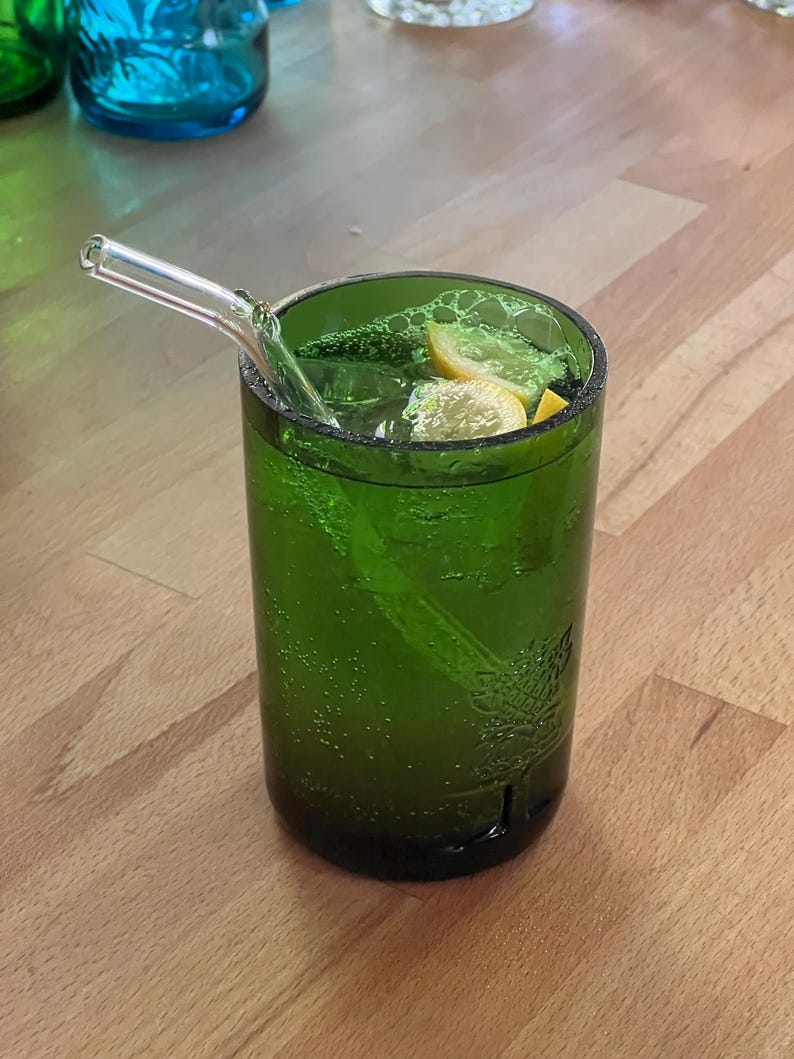 May include: A green glass tumbler filled with a green beverage, ice, and lemon slices. A clear glass straw extends from the drink. The glass has a textured surface and is sitting on a wooden surface.