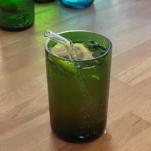 May include: A green glass with a refreshing green beverage, a lemon slice, and mint leaves. A clear glass straw is inserted into the drink. Other green and blue glass bottles are in the background.