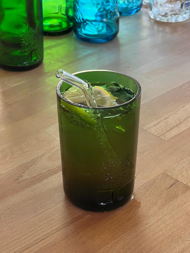 May include: A green glass with a refreshing green beverage, a lemon slice, and mint leaves. A clear glass straw is inserted into the drink. Other green and blue glass bottles are in the background.