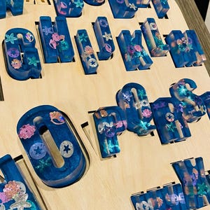Close-up macro shot of the smooth wooden puzzle insert and a chunky blue glitter resin letter with embedded seashell shapes.