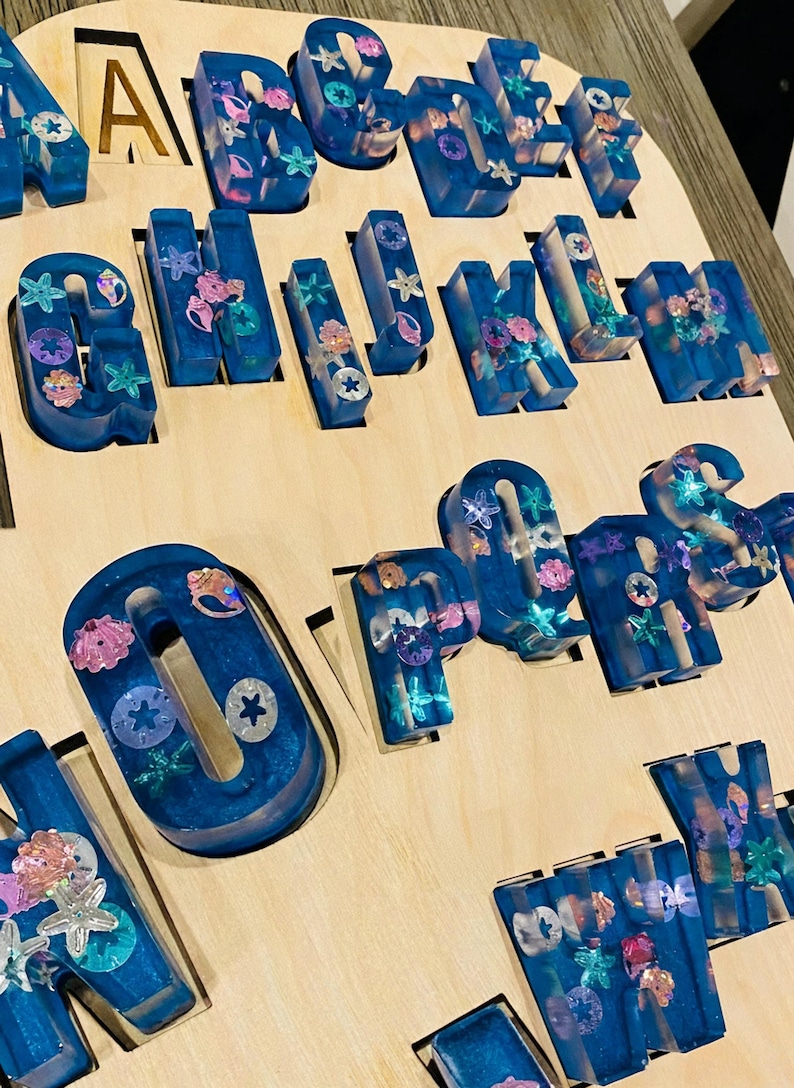 Close-up macro shot of the smooth wooden puzzle insert and a chunky blue glitter resin letter with embedded seashell shapes.