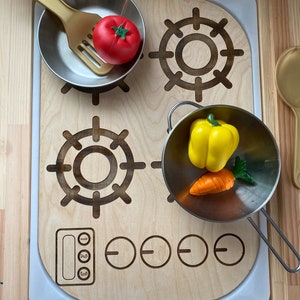 Puede incluir: Una estufa de cocina de juguete de madera con dos ollas plateadas, un tomate rojo, un pimiento amarillo, una zanahoria naranja y una espátula dorada. La estufa tiene cuatro perillas y un panel de control con tres botones.