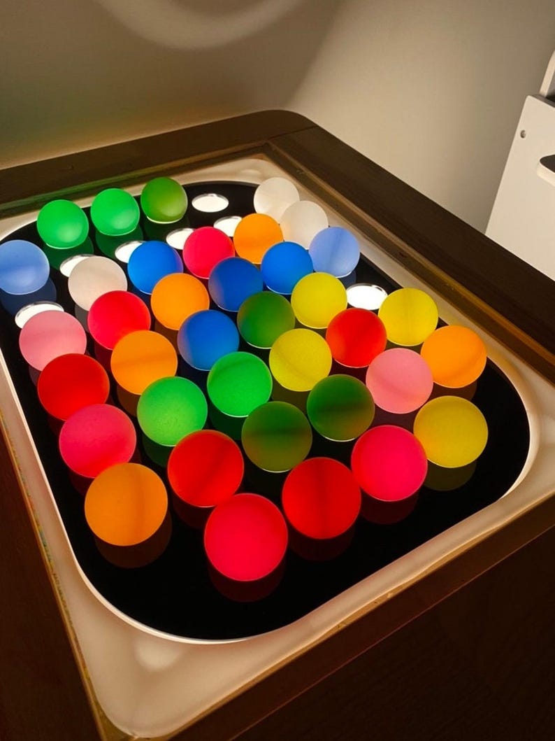 A black acrylic insert with 32 colorful, translucent ping pong balls, lit from below, in a light-colored wooden table.