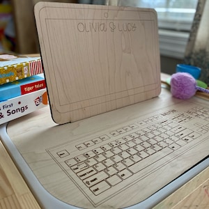 Pode incluir: Uma secretária infantil de madeira clara com um design de teclado e um painel traseiro em forma de tablet. O painel traseiro é gravado com os nomes "Olivia & Lucy".