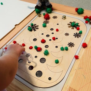 Puede incluir: Un tablero de actividades de madera con forma de árbol de Navidad con círculos y formas de media luna recortados. El tablero está rodeado de pompones rojos y verdes y copos de nieve. Una mano de niño está usando una herramienta para colocar los pompones en los agujeros.