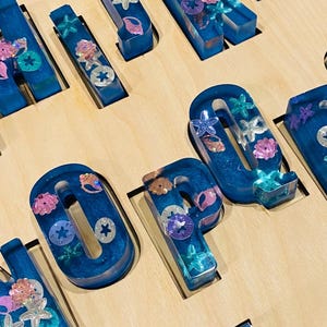 A close-up of a wooden puzzle board, showing several chunky, translucent blue resin letters with embedded glitter and seashell shapes, fitting into the board's cutouts.